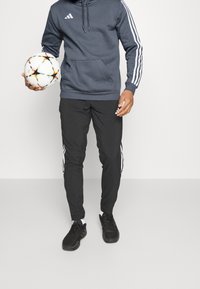 Felpa grigia scura con strisce bianche, pantaloni sportivi neri con dettagli bianchi e scarpe da ginnastica nere. Tiene un pallone da calcio multicolore.