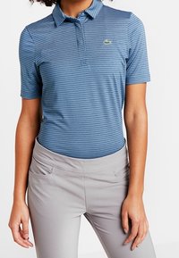 Person som bär en blårandig kortärmad pikétröja och ljusgrå byxor, som står mot en plain vit bakgrund.