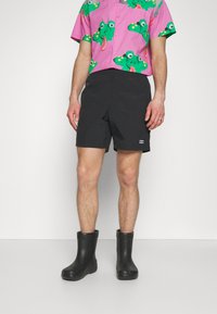 Calças curtas pretas com uma textura suave, combinadas com uma camisa rosa estampada com um padrão de rã colorido e botas de borracha pretas. Notável pelos contrastes de cores e design divertido.