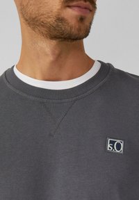 Sudadera gris con cuello redondo, puños acanalados y un pequeño logo bordado en el pecho. Tejido suave con una textura sutil.