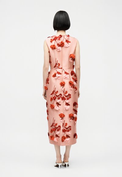 Lühikeste mustade juustega naine, kes kannab varrukateta roosat midi-kleiti, mis on kaunistatud punaste ja roosade lilleaplikatsioonidega, ning valget värvi kontsakingi.