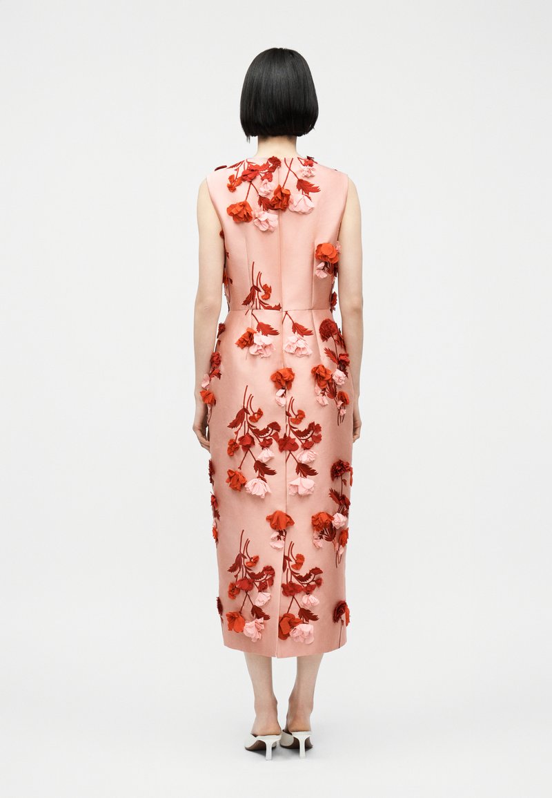 Donna con capelli corti neri che indossa un vestito midi rosa senza maniche decorato con applicazioni floreali rosse e rosa, abbinato a sandali bianchi con tacco.