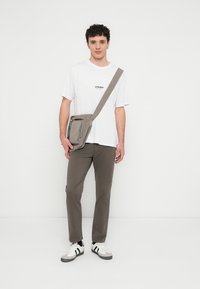 Τσάντα crossbody από ανοιχτό γκρι ύφασμα με φερμουάρ σε εμπρόσθια τσέπη, φορεμένη στον ώμο, σε συνδυασμό με λευκό t-shirt και χακί παντελόνι.