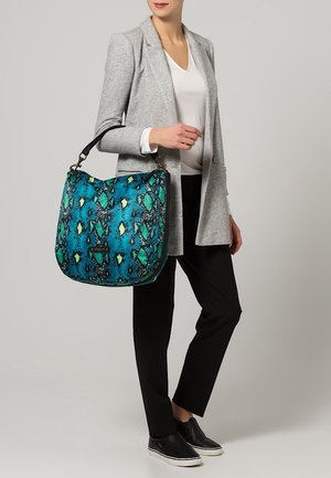Handtasche mit blauem und grünem Schlangenmuster, aus strukturiertem Stoff, mit einem schwarzen Tragegurt und Metallbeschlägen, von einer Person getragen.