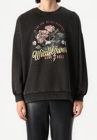 Sudadera negra de gran tamaño con gráfica floral y texto "Wildflower Rock & Roll" en dorado y rosa. Puños y dobladillo acanalados. Material de mezcla de algodón.