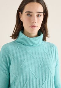 Pull en turtleneck turquoise avec texture côtelée et motifs angulaires. Tissu doux, design ajusté et col repliable bien en vue.
