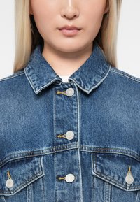 Jeans jakna u srednje plavoj boji s klasičnom kragnom, srebrnim dugmadi i džepovima s preklopom. Ima teksturiranu površinu i vidljive detalje šivanja.