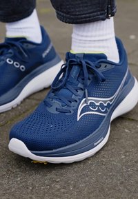 Navy blue athletic shoe with mesh upper, white accents, and rounded sole. Features lace-up design with branding and textured surfaces.
