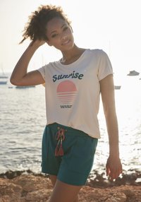 Camiseta beige clara con texto verde "Sorpresa" y gráfico de un sol rosa. Pantalones cortos teal oscuro con bolsillos laterales y cordón con borlas rosas.