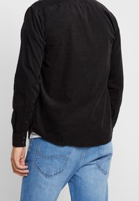 Camisa negra de manga larga con acabado texturizado y puños con botones, combinada con vaqueros azul claro con bolsillos traseros curvados.