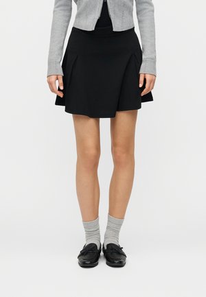 Donna con una minigonna plissettata nera, cardigan grigio, calzini grigi e mocassini neri, in piedi su uno sfondo bianco semplice.