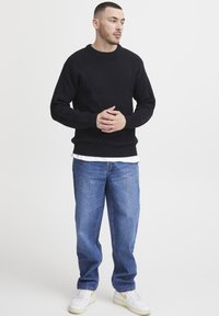 Man with short hair wearing a black sweater, blue jeans, and white sneakers standing clasping hands, looking to the side on white background.