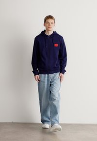 Marineblå zip-up hettegensere med to frontlommer, en liten rød label på brystet, kombinert med løse lyseblå jeans og hvite sneakers.