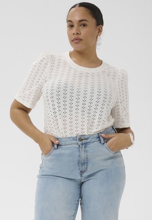 Vrouw draagt een wit eyelet topje met pofmouwen, ingestopt in lichtblauwe, hooggetailleerde jeans, handen in de zakken, tegen een effen achtergrond.