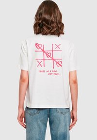 Maglietta bianca in cotone a maniche corte, con una grafica di tris rosa e la scritta "TRE IN FILA NON IL TUO..." sul retro.
