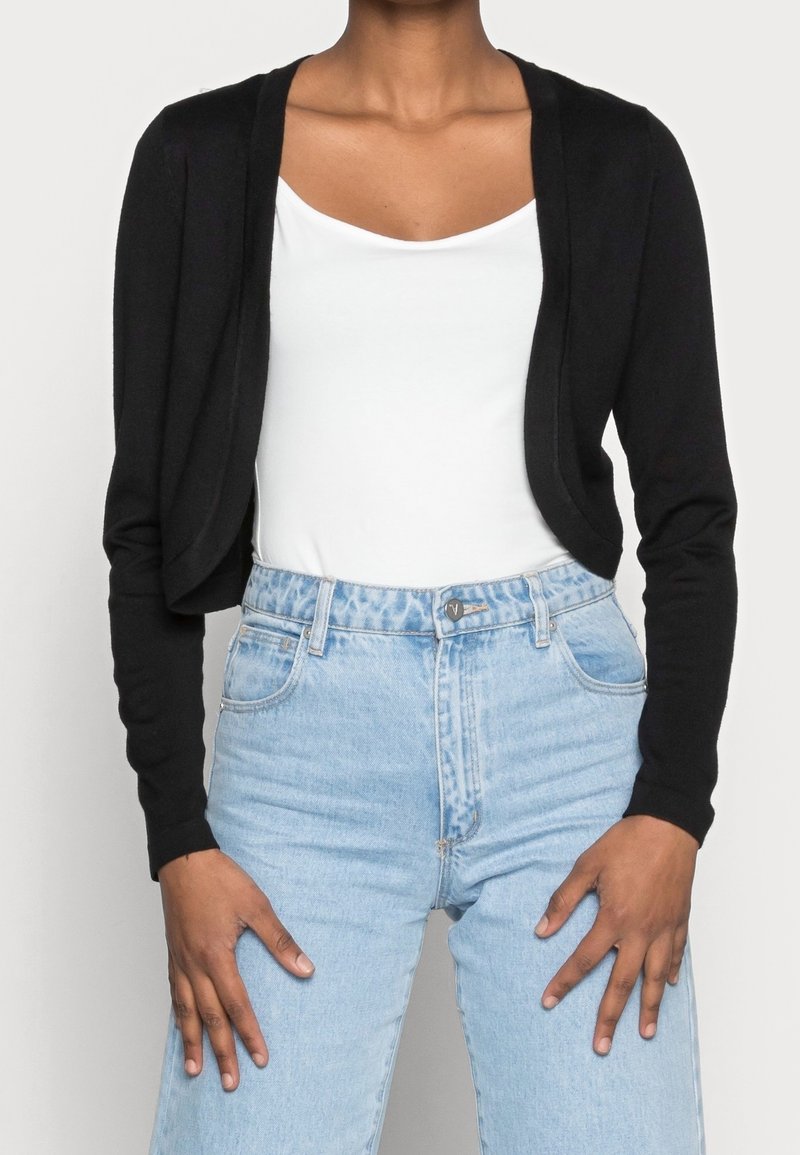 Svart kort cardigan i mjukt tyg, med långa ärmar och öppen front, som bärs tillsammans med en vit t-shirt och ljusblå högmidjade jeans.