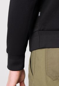 Sweatshirt noir avec des poignets texturés et une coupe ajustée. Le matériau semble doux, associé à un pantalon vert olive avec des poches visibles.
