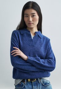 Junge Frau mit langen dunklen Haaren, die einen blauen Strickpullover und Jeans trägt, steht mit gekreuzten Armen vor einem schlichten hellen Hintergrund.