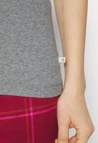 Grå ribbet tanktop med avrundet kant, med et lite stoffmerke på siden, brukt med et rødt og rosa rutete skjørt.