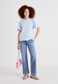 T-shirt en coton bleu clair avec de courtes manches bouffantes, associé à un jean en denim taille haute. Le mannequin tient un petit sac à main rose avec une bandoulière en chaîne.