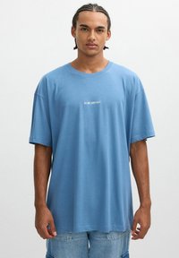 Camiseta azul de gran tamaño hecha de algodón suave. Presenta un escote redondo y mangas cortas. Texto "24 HORAS DE SERVICIO" impreso en el pecho.
