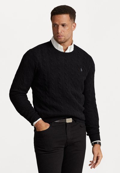 Schwarzer Strickpullover mit gerippten Bündchen, kombiniert mit einem weißen Hemd mit Kragen und schwarzen Hosen. Der Look wird mit einer metallischen Gürtelschnalle abgerundet.