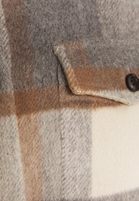 Ullrutigt tyg i grått, brunt och creme med en närbild av en knapp och en sydd fickdetalj. Mjuk textur med ett varmt utseende.