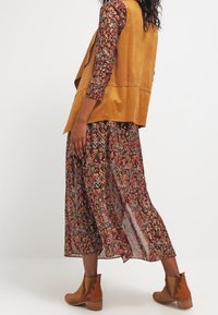 Hnědá semišová vesta se cvočky přes vícebarevné vzorované maxi šaty. Outfit doplňují hnědé kotníkové boty.