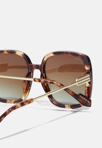 Occhiali da sole oversize in acetato tartarugato con lenti marroni sfumate e dettagli in metallo color oro. Presentano una montatura quadrata audace e il logo del marchio sulla tempia.