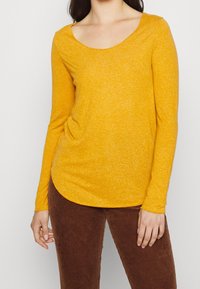 Långärmad t-shirt med rundad halsringning i senapsgul färg, tillverkad av textil med en subtil melerad mönstring, kombinerad med bruna manchesterbyxor.