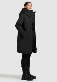 Nais, kes kannab pikka musta kapuutsiga talvehudjat, käed taskutes, mustad retuusid ja mustad paksud saapad, seisab külgliikudes halli tausta ees.