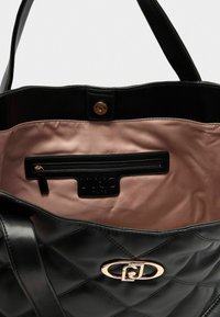 Zwarte gewatteerde tote bag met een glanzende afwerking, twee handvatten, een roze satijnen binnenkant en een ritsvak met een logo-label. Vergulde accenten.