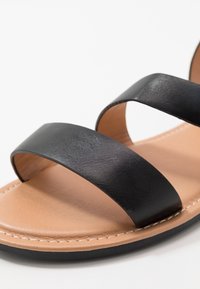 Mustat nahkaiset sandaalit, joissa on kaksi leveää nauhaa. Pohja on tan-värinen, sileäpintainen ja reunassa näkyy ommel.