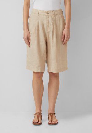Donna che indossa pantaloncini beige in lino fino al ginocchio, top bianco senza maniche e sandali bassi marroni su uno sfondo chiaro e uniforme.