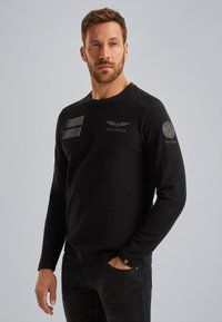Schwarzes Langarmshirt aus weichem Material mit geprägten Patches auf der Brust und dem Ärmel, runder Ausschnitt und taillierte Passform.
