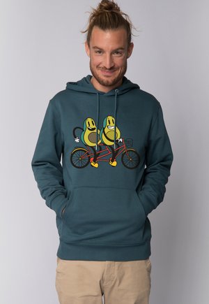Mann mit einem Dutt, der lächelt und einen türkisfarbenen Hoodie trägt, der zwei lächelnde Avocados zeigt, die gemeinsam auf einem Tandemfahrrad fahren.