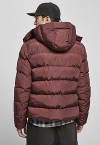Bordo puffer jakna s kapuljačom, kviltani dizajn, glatka tekstura i vidljive šavove, nosi se preko košulje s kariranim uzorkom.