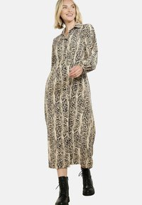 Žena se usmívá, má na sobě dlouhé šaty s hadím potiskem na knoflíky s límečkem a černé šněrovací kotníkové boty, jde proti jednobarevnému pozadí.