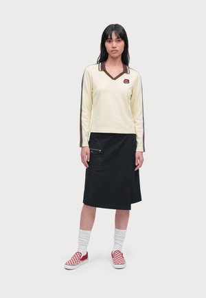 Femme portant un haut crème à manches longues avec un col marron et des rayures, une jupe noire longueur genoux, des chaussettes blanches et des baskets rouges à carreaux sans lacets.