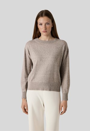 Kobieta z długimi brązowymi włosami ubrana w beżowy dziergany sweter i białe spodnie, stojąca na jednolitym szarym tle.