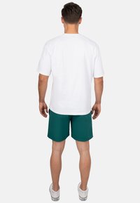 T-shirt bianco in cotone con maniche corte, abbinato a pantaloni corti color teal, indossato con scarpe da ginnastica bianche. Il tessuto appare liscio e rilassato.