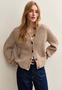 Beige stickad cardigan med ribbad textur, rund halsringning och bruna knappar. Buren över en vit topp, som matchar med mörk Denim jeans.
