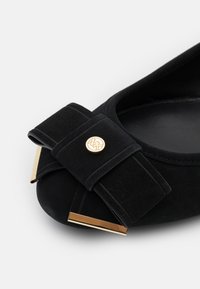 Ballerine en cuir suédé noir avec un nœud décoratif, des finitions en métal doré et un bout arrondi. Surface texturée avec des accents de couture contrastés.