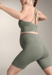Olivnozeleni športni modrček z vijugastimi naramnicami in ujemajočimi se kratkimi hlačami z visokim pasom, izdelan iz gladkega, raztegljivega materiala.