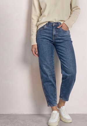 Frau in blauen High-Waist-Jeans, cremefarbenem Pullover und weißen Sneakers steht mit einer Hand in der Tasche vor einer schlichten Wand.