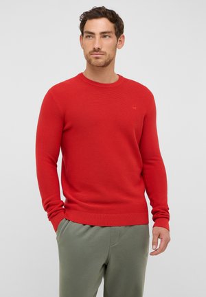 Czerwony sweter dziany z okrągłym dekoltem i ściągaczami, połączony z jasnochłodnymi spodniami. Subtelny detal z logo na klatce piersiowej.