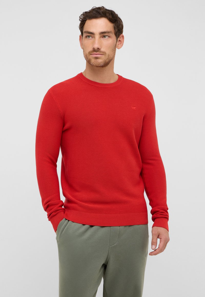 Roter, gestrickter Pullover mit Rundhalsausschnitt und gerippten Bündchen, kombiniert mit hellgrünen Hosen. Subtiler Logo-Detail auf der Brust.