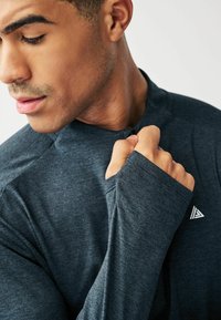 Dunkelgraues sportliches Langarmshirt mit hohem Kragen, taillierten Ärmeln und einer subtilen Textur. Verfügt über ein kleines dreieckiges Logo auf der Brust.