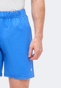 Άνδρας που φοράει αθλητικές σορτς με φωτεινό μπλε ελαστική μέση και λευκό μπλουζάκι, το χέρι χαλαρό στο πλάι.