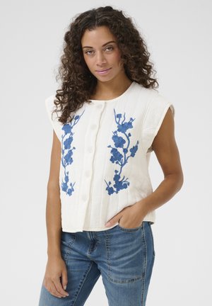 White sleeveless vest with blue floral embroidery, featuring button closure and textured fabric. Worn over blue denim jeans.